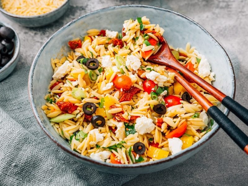 Eine blaue Schale mit dem Reisnudelsalat auf einem grauen Untergrund. Links daneben eine Schale mit rohen Reisnudeln und eine kleine Schale mit schwarzen Oliven.