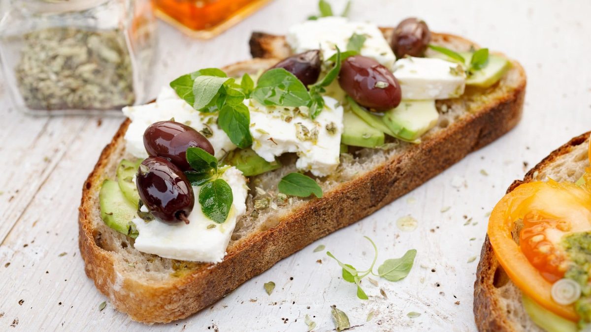 Draufsicht: Ein griechisches Sandwich mit Oliven und Feta und Oregano liegt auf einem weißen Holztisch.