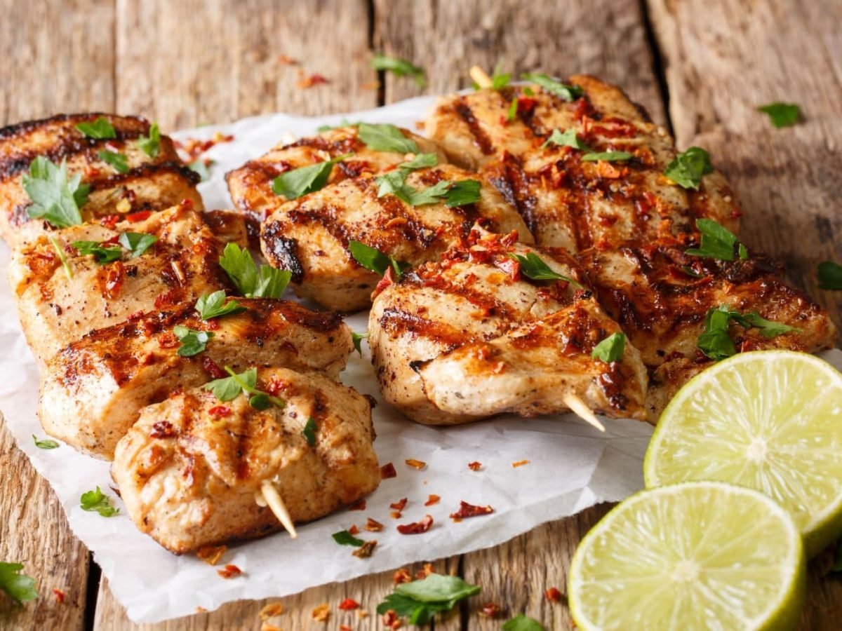 Zwei Hähnchenspieße in Joghurt-Kräuter-Marinade mit frischen Kräutern auf weißem Teller auf Holt. Im Bild eine halbierte Zitrone. Draufsicht.