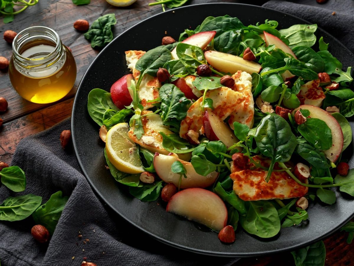 Ein schwarzer Teller mit dem Halloumi-Salat auf einem dunklen Holztisch mit Spinat und Haselnüssen.