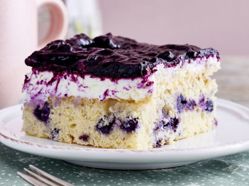 Ein Stück von einem Heidelbeer-Quarkkuchen auf einem weißen Teller von vorne fotografiert auf einer grünen Tischdecke mit weißen Punkten. Hinten links eine rosarote Tasse.