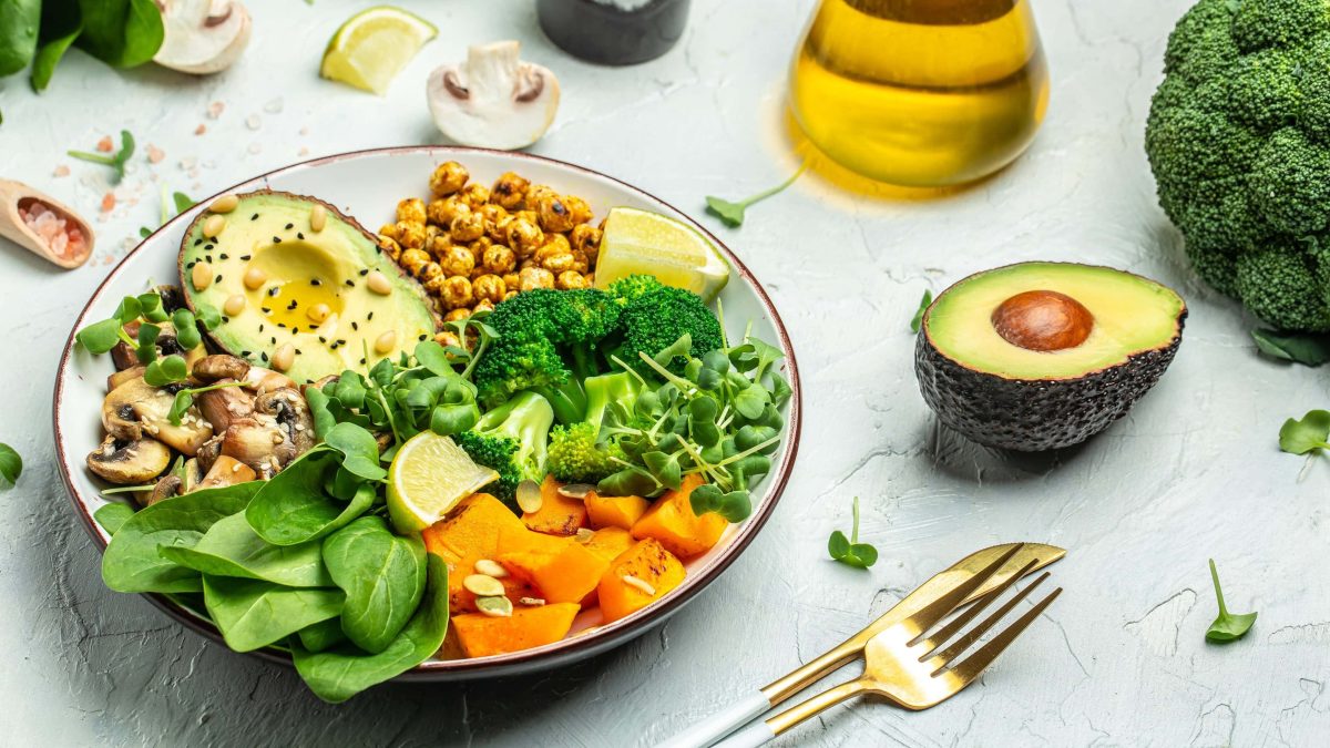 Eine herzhafte Frühstücksbowl mit Kichererbsen, Süßkartoffel, Spinat, Avocado, Brokkoli, Pilzen. Daneben goldenes Besteck, eine halbe Avocado, eine Flasche Öl und ein Brokkoli.