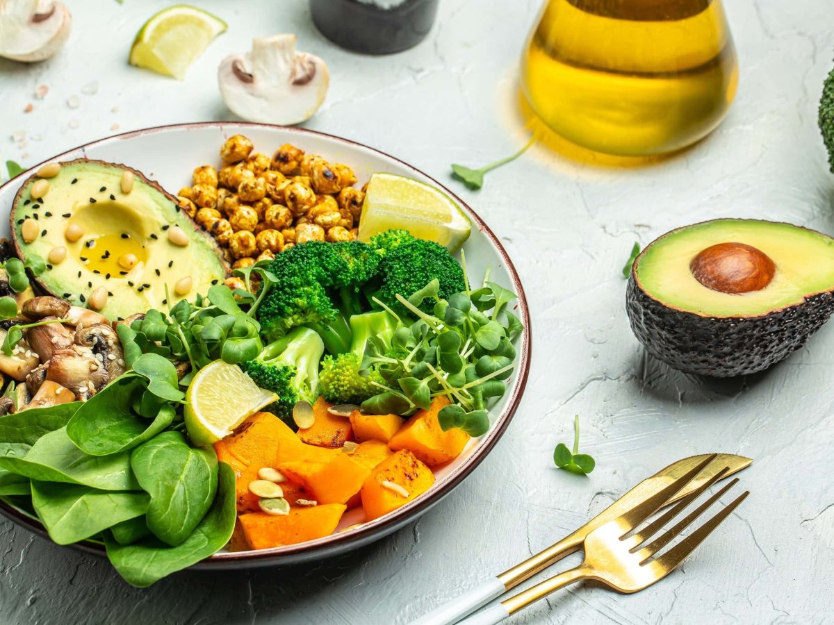 Eine herzhafte Frühstücksbowl mit Kichererbsen, Süßkartoffel, Spinat, Avocado, Brokkoli, Pilzen. Daneben goldenes Besteck, eine halbe Avocado, eine Flasche Öl und ein Brokkoli.