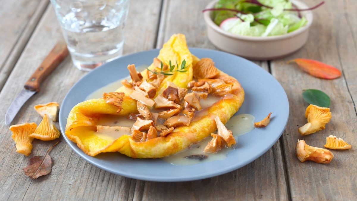 ein Pfannkuchen mit Pfifferlingen und Rahmsauce gefüllt, drumherum frische Pilze, ein Glas Wasser, ein Messer, eine Schüssel Salat und bunte Blätter.