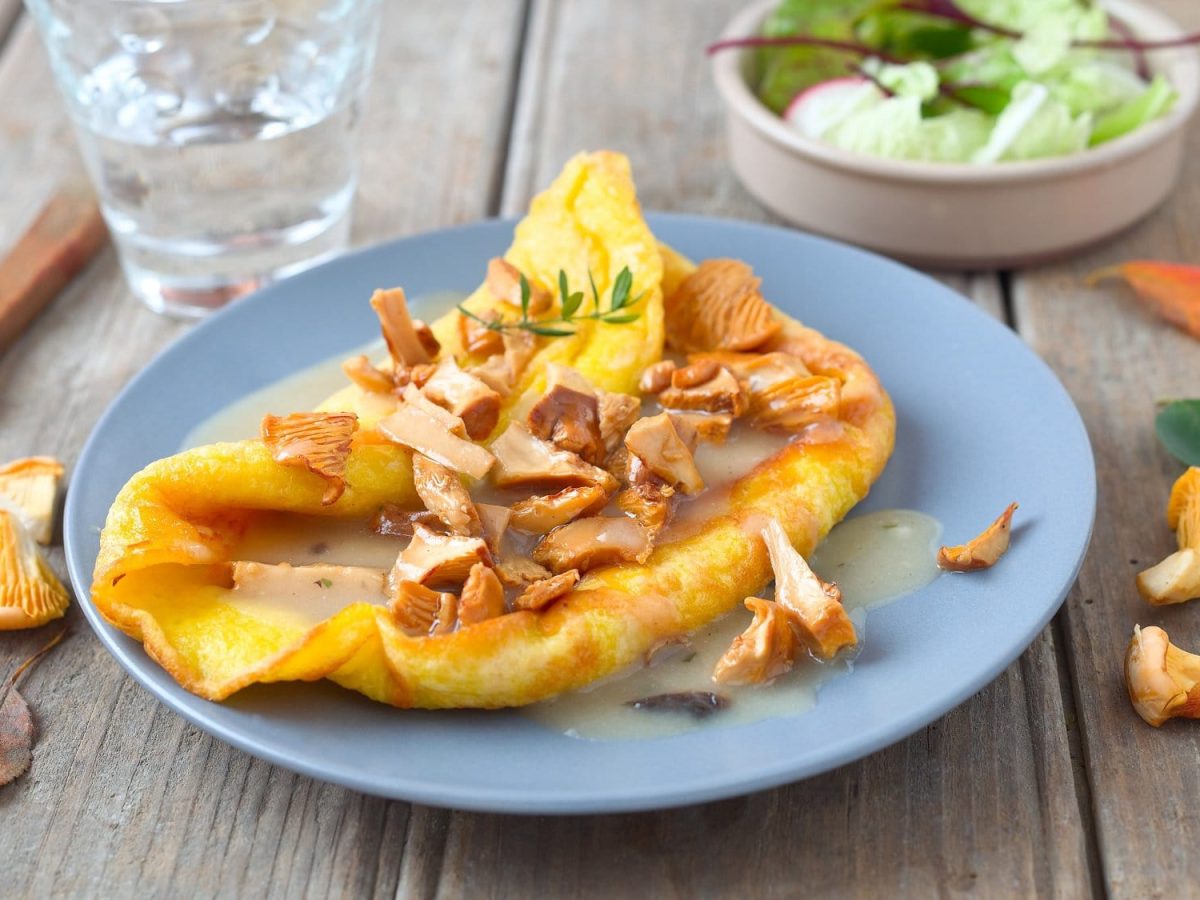 ein Pfannkuchen mit Pfifferlingen und Rahmsauce gefüllt, drumherum frische Pilze, ein Glas Wasser, ein Messer, eine Schüssel Salat und bunte Blätter.