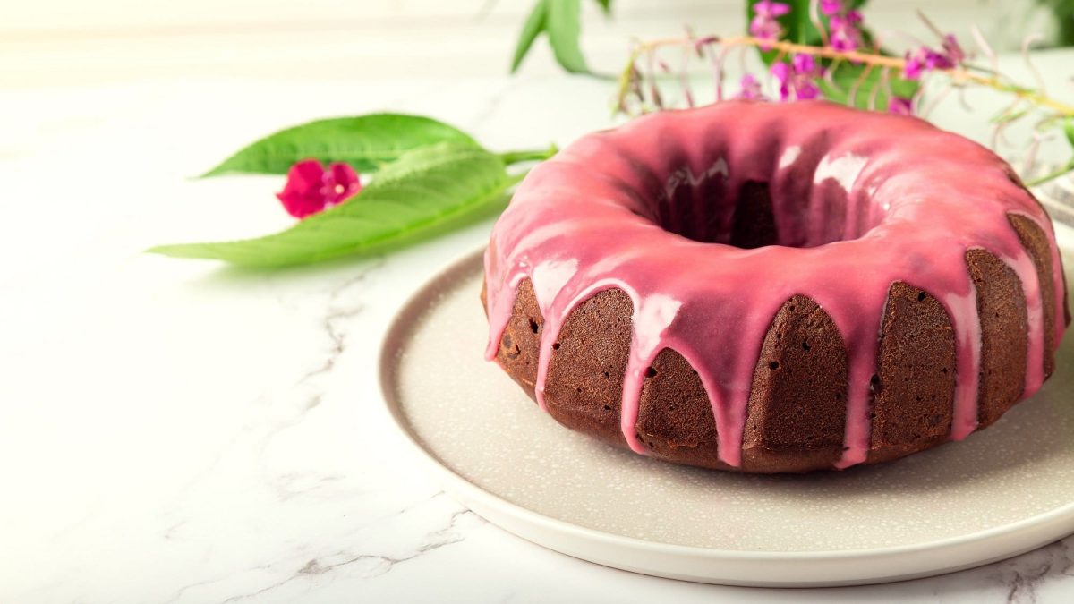 Ein Himbeer-Gugelhupf mit weißer Schokolade und rosa Glasur auf einem Teller, dahinter liegen rosablühende Pflanzen.