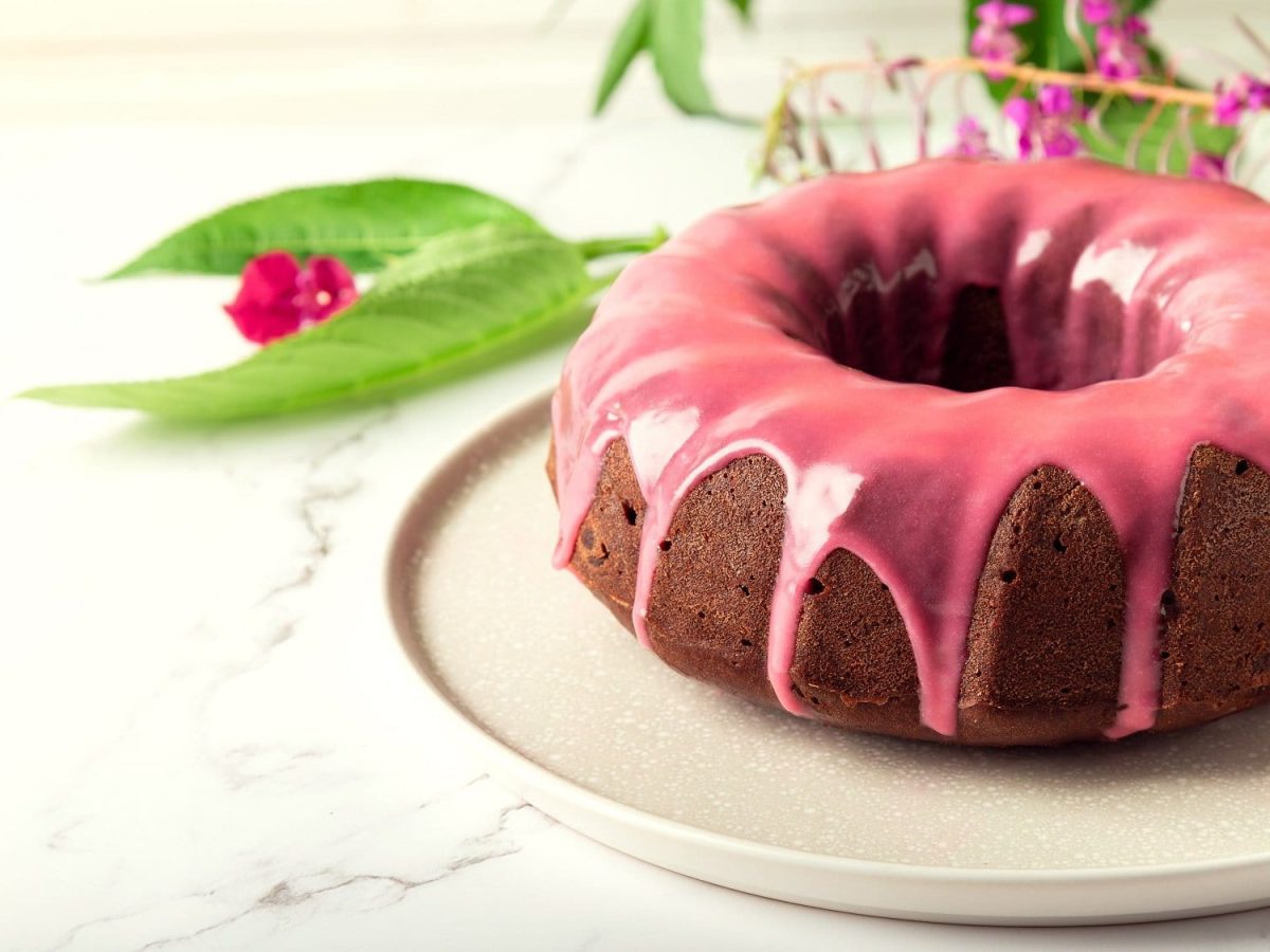 Ein Himbeer-Gugelhupf mit weißer Schokolade und rosa Glasur auf einem Teller, dahinter liegen rosablühende Pflanzen.