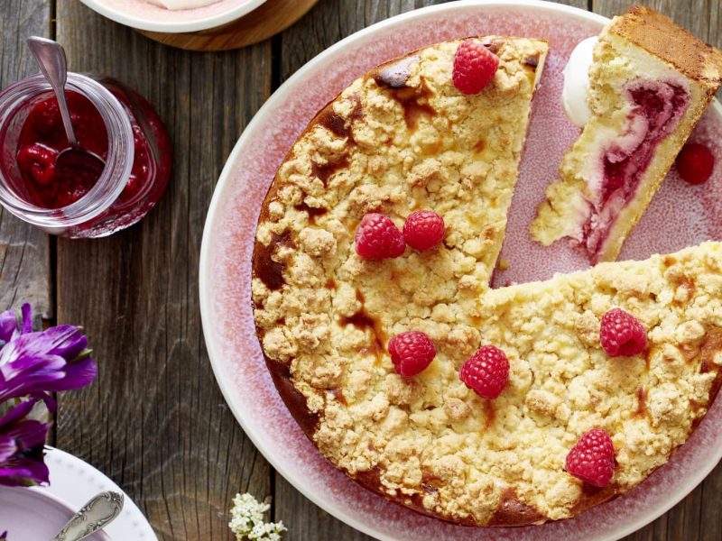 Ein Himbeer-Käsekuchen auf einem hellroten Servierteller auf einem hölzernen Hintergrund. Links davon ein Glas gefüllt mit Himbeerkompott und Blumen.