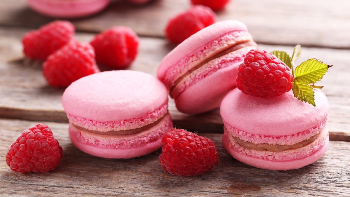 Frontalsicht: drei Himbeer-Macarons liegen auf einem Untergrund aus Holz. Drumherum liegen frische Himbeeren.