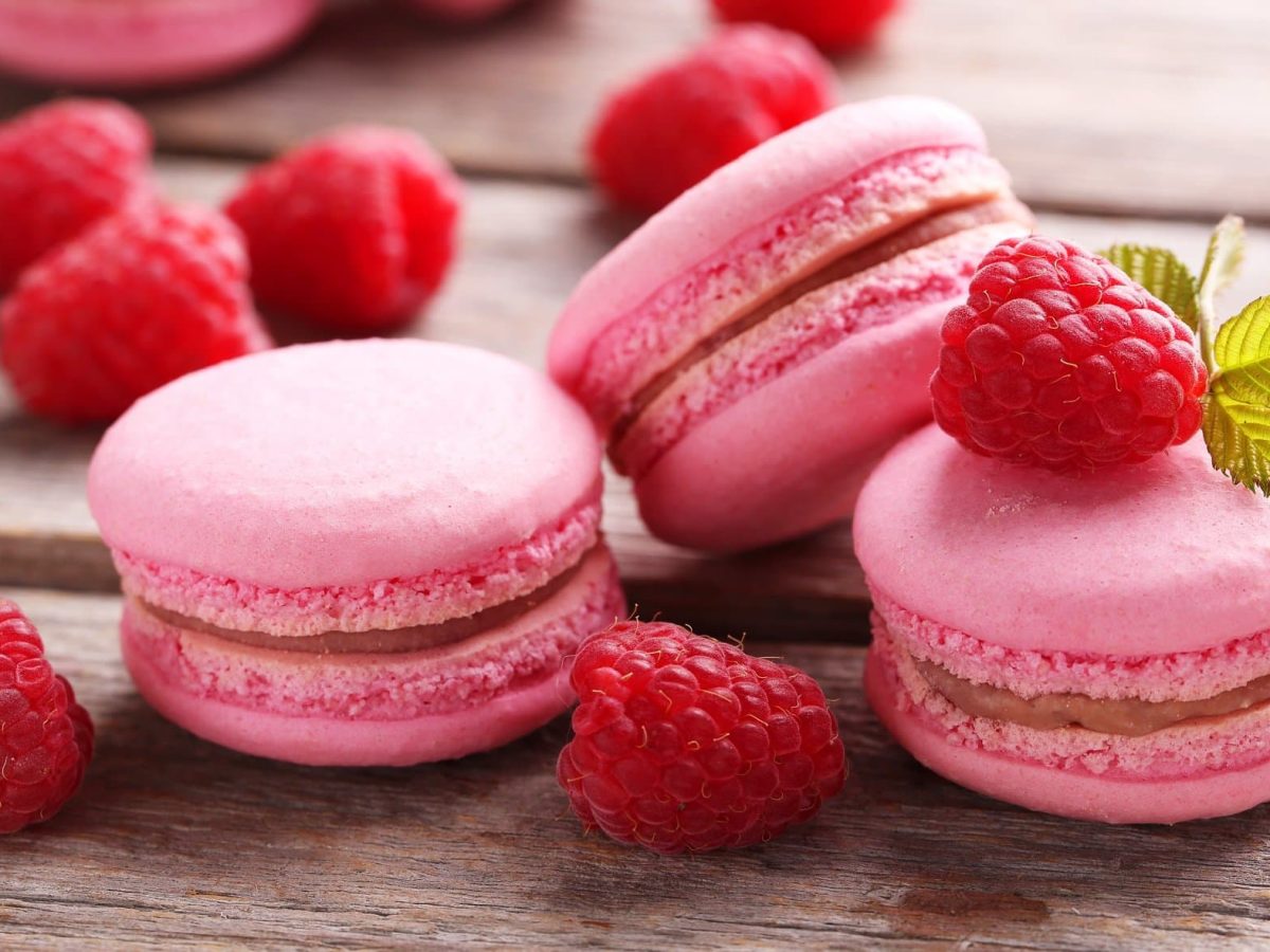 Frontalsicht: drei Himbeer-Macarons liegen auf einem Untergrund aus Holz. Drumherum liegen frische Himbeeren.