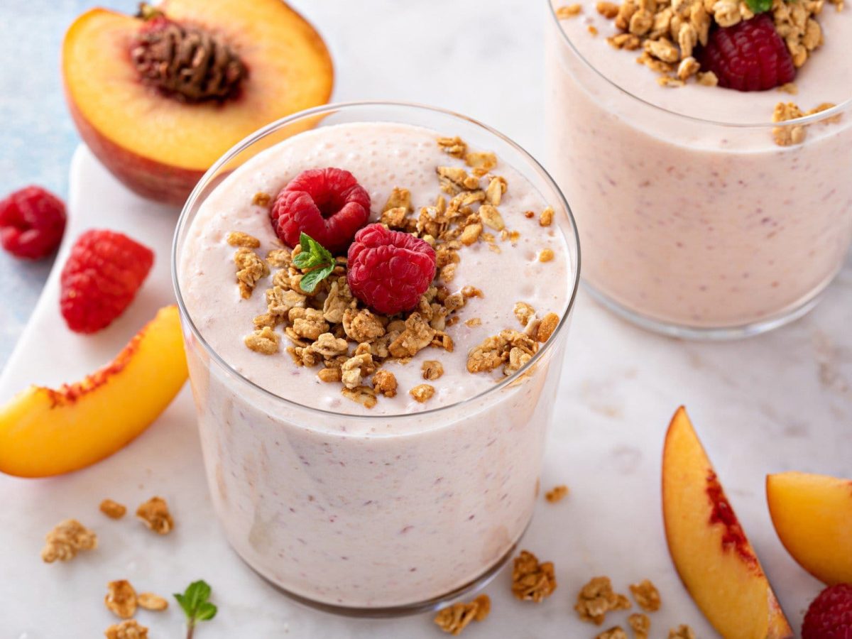 Draufsicht: Zwei Gläser Himbeer-Pfirsich-Smoothie. Daneben liegen Pfirsich-Spalten, Himbeeren und Granola.