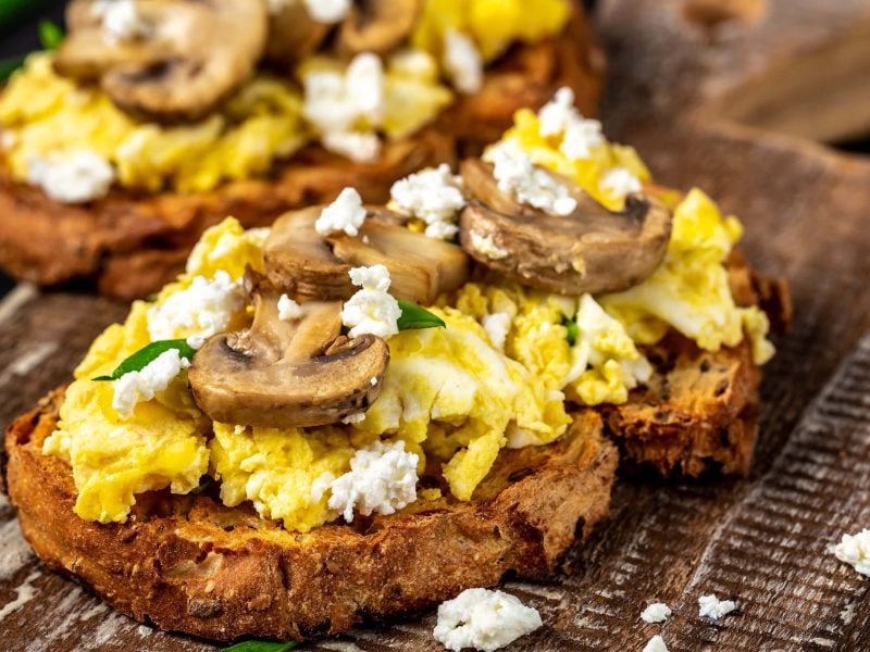 Zwei Scheiben mit dem Eiweißbrot und Hüttenkäse-Rührei auf einem dunklen Holzbrett.