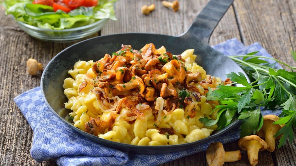 Selbst gemachte Käsespätzle mit Pfifferlingen und Röstzwiebeln und Petersilie in schwarzer Gusspfanne auf blauem Tuch und Holz. Daneben frische Pfifferlinge, Petersilie und einer Glasschüssel mit grünem Salat. Draufsicht.