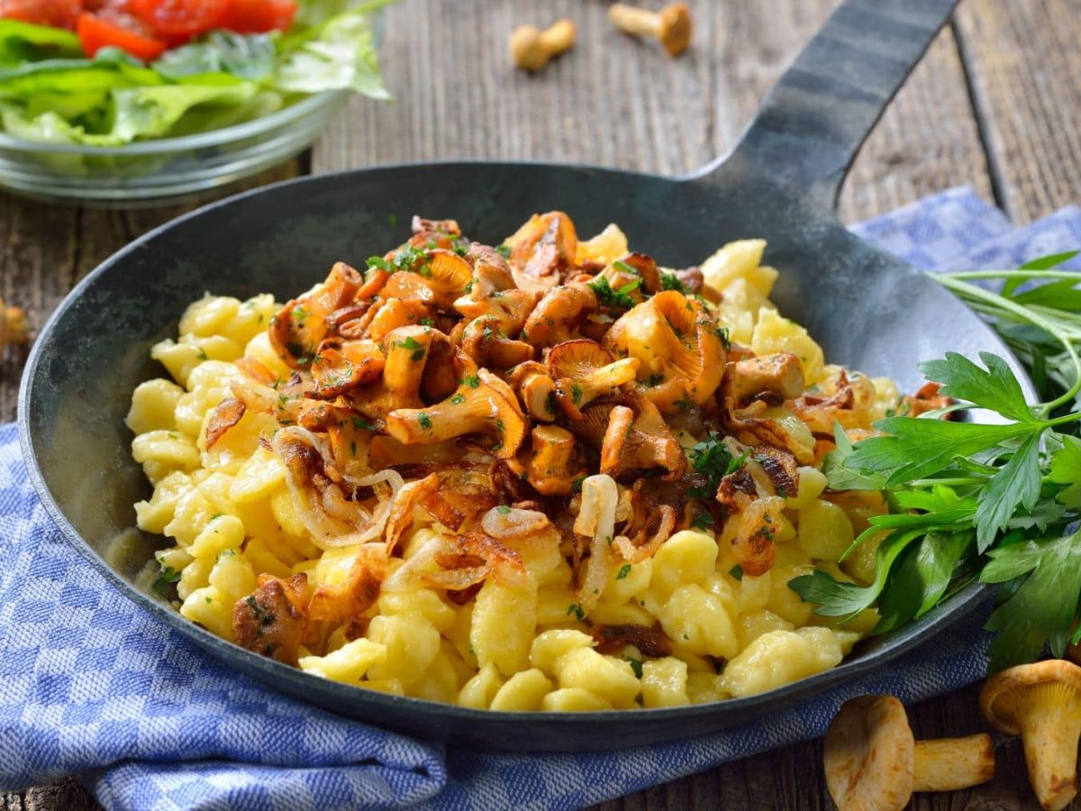 Selbst gemachte Käsespätzle mit Pfifferlingen und Röstzwiebeln und Petersilie in schwarzer Gusspfanne auf blauem Tuch und Holz. Daneben frische Pfifferlinge, Petersilie und einer Glasschüssel mit grünem Salat. Draufsicht.