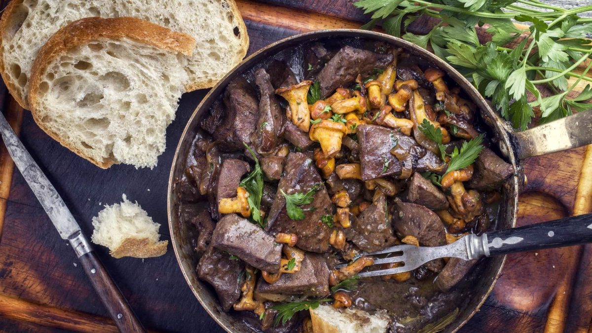 Draufsicht: Auf einem Untergrund aus Holz liegt eine dunkle Pfanne mit Kalbsleber mit Pfifferlingen. Drum herum liegen frische Petersilie sowie zwei Scheiben Weißbrot.