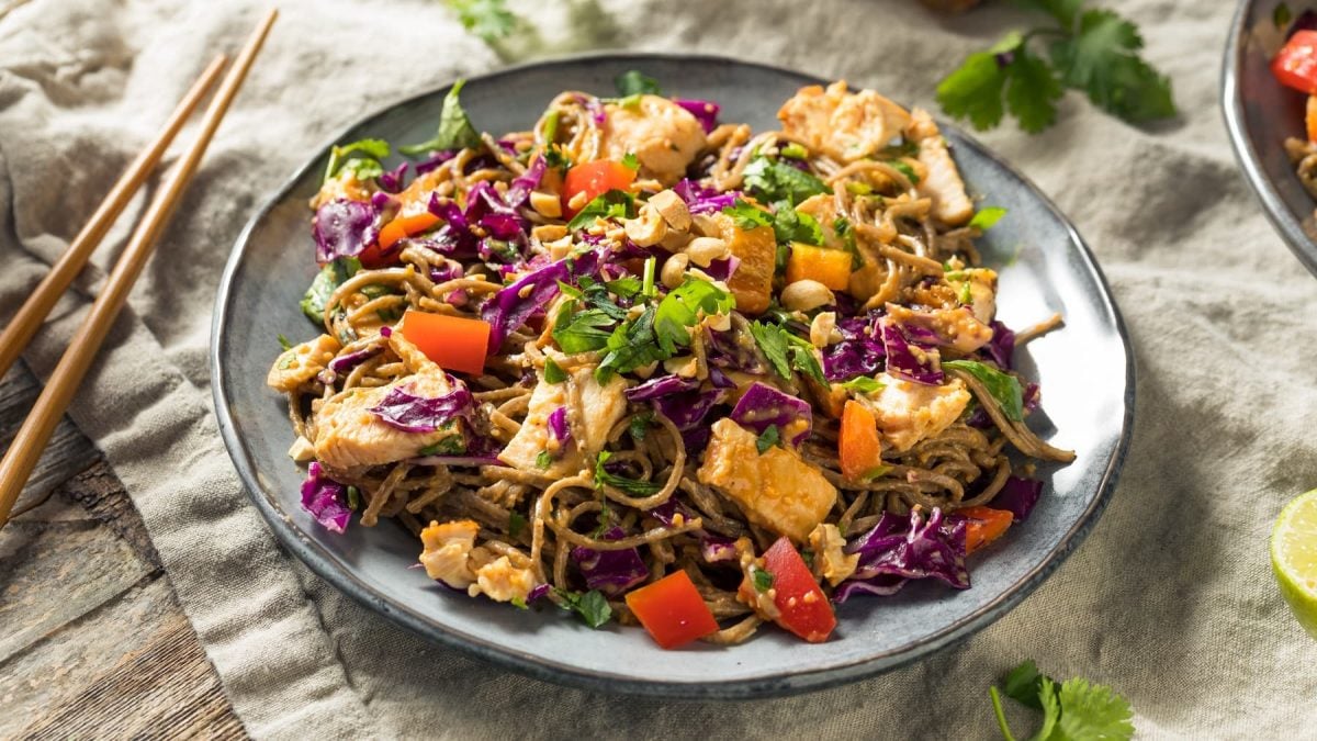 Ein grauer Teller mit den kalten Soba Nudeln und Erdnusssauce. Der Teller steht auf einem Holztisch mit einer grauen Tischdecke, Stäbchen und Koriander.