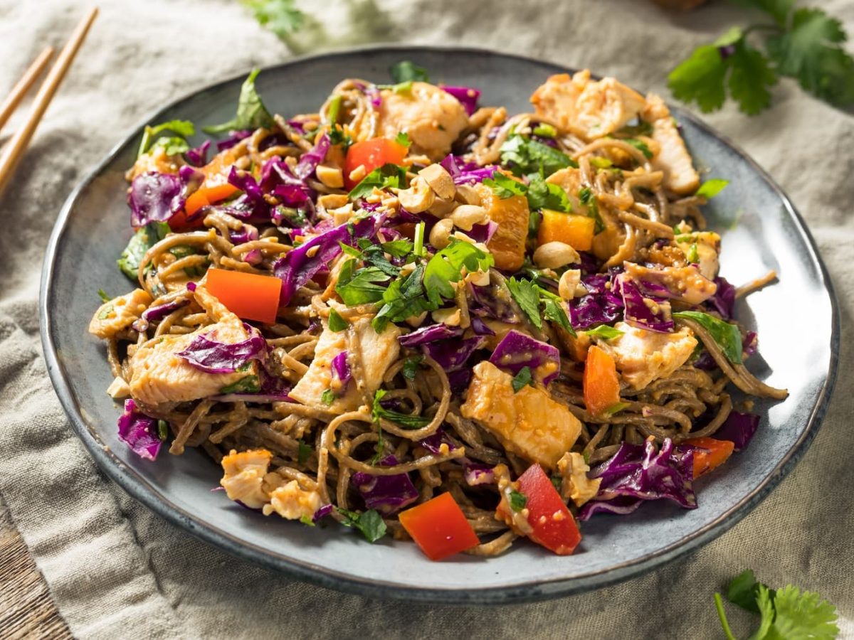 Ein grauer Teller mit den kalten Soba Nudeln und Erdnusssauce. Der Teller steht auf einem Holztisch mit einer grauen Tischdecke, Stäbchen und Koriander.