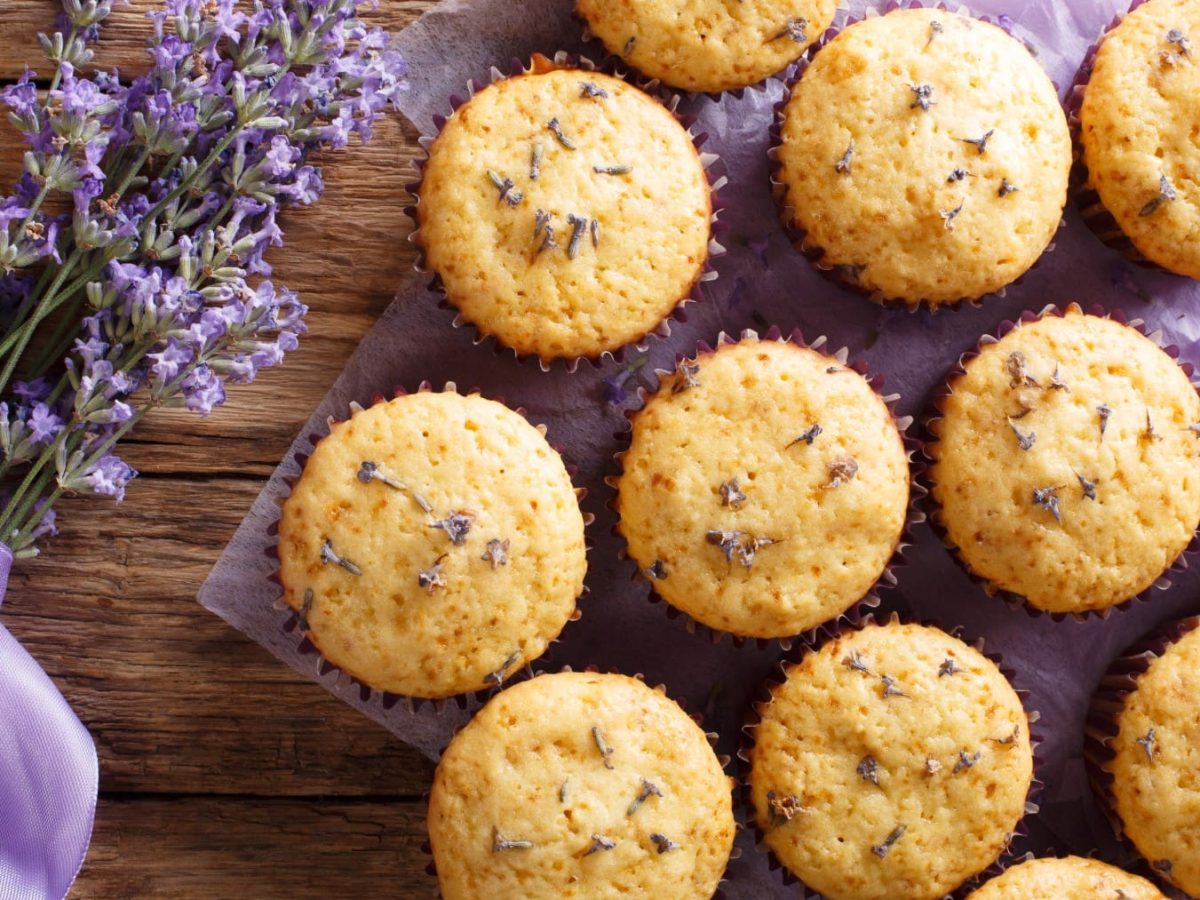 Draufsicht: Mehrere Lavendel-Muffins auf einem lila Tuch, daneben ein Bund Lavendel, Holzuntergrund.