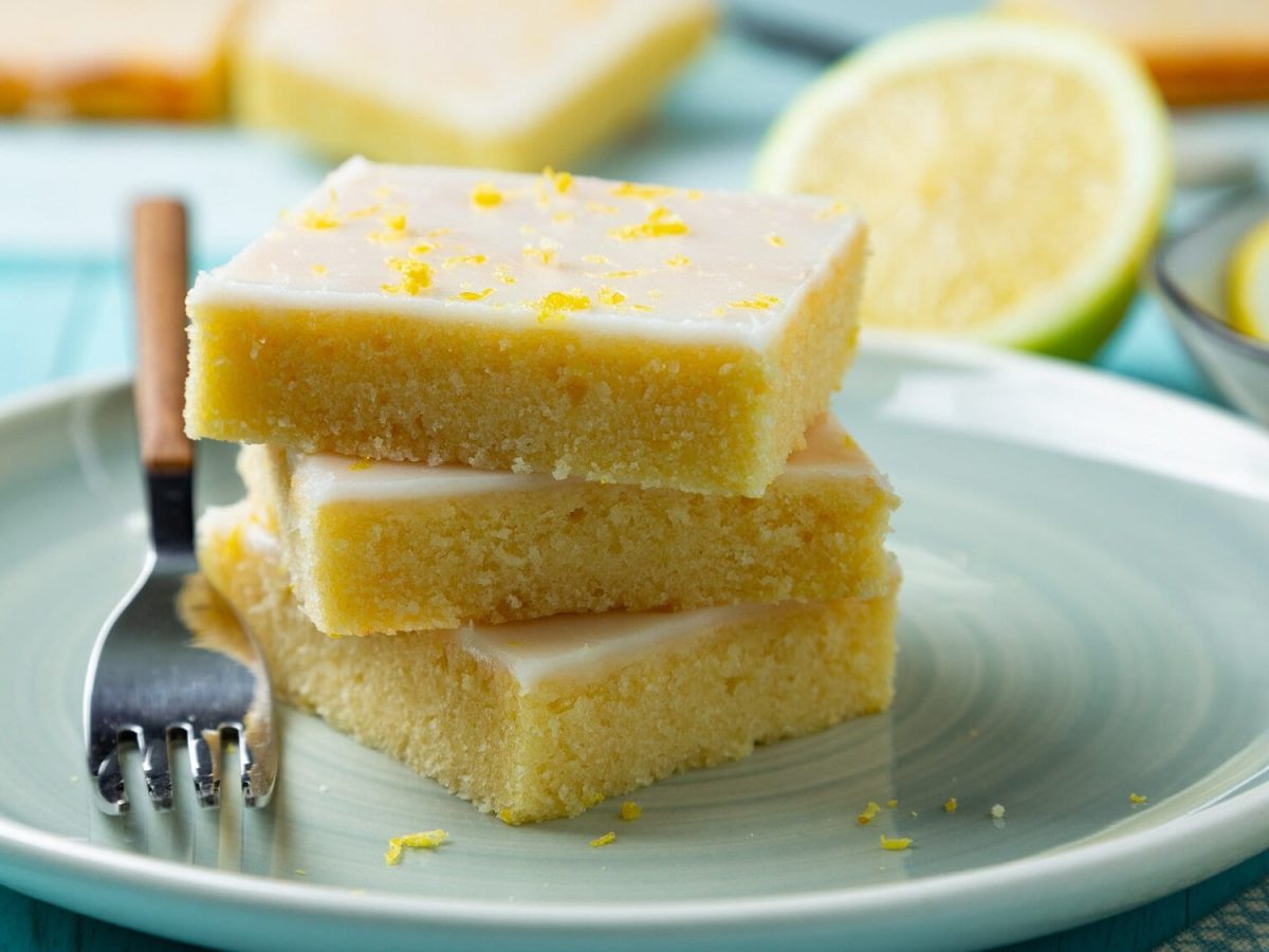 Drei gestapelte Stücke Lemonies, Zitronen-Brownies mit Zuckerguss, auf einem Teller, auf dem auch eine Gabel liegt. Dahinter ein paar Zitronen.