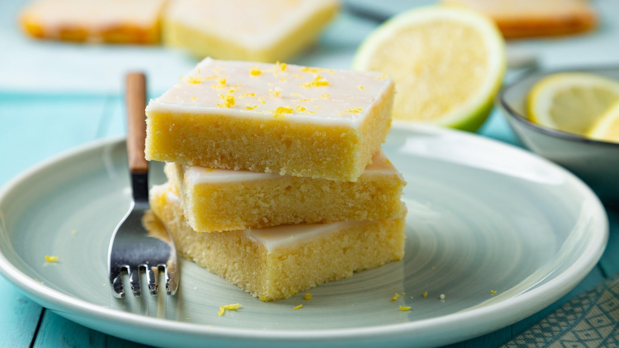 Drei gestapelte Stücke Lemonies, Zitronen-Brownies mit Zuckerguss, auf einem Teller, auf dem auch eine Gabel liegt. Dahinter ein paar Zitronen.