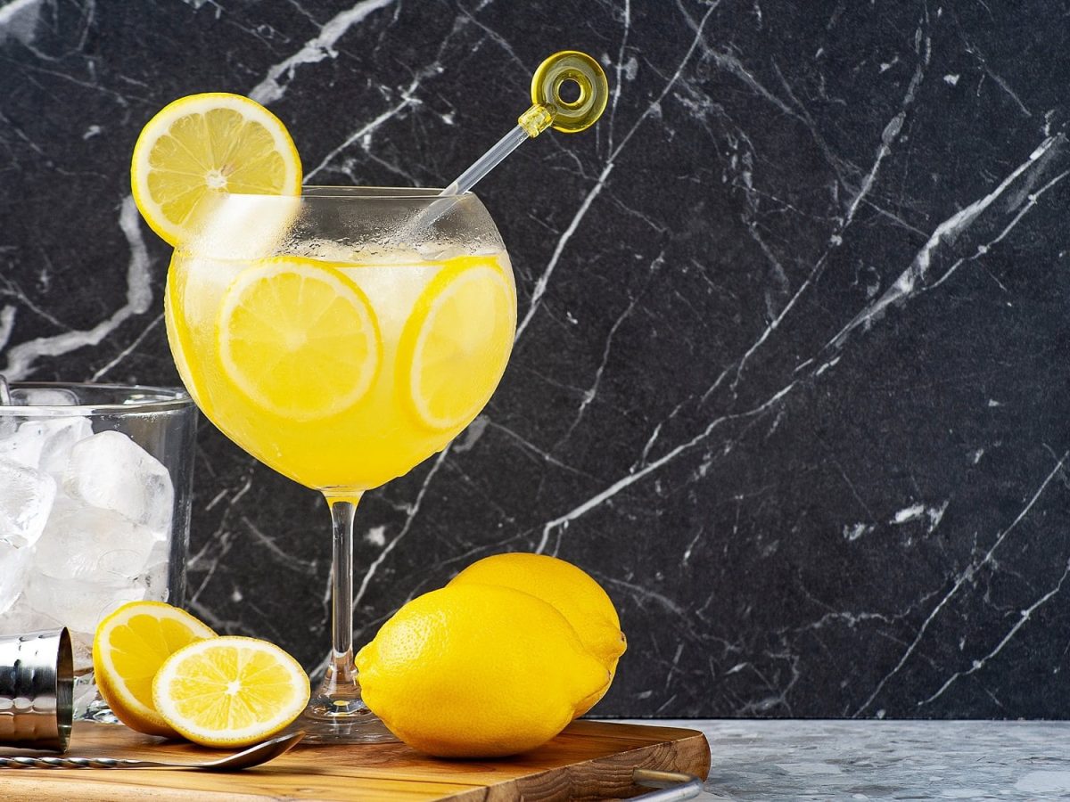 Ein Glas Limoncello Gin Fizz mit Eiswürfeln und Zitronenscheiben auf einem Brett, darauf auch frische Zitronen und ein Glas mit Eiswürfeln.