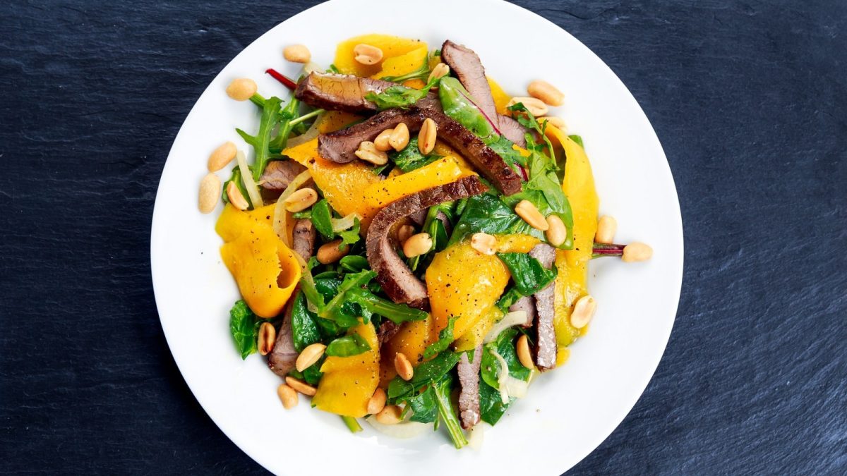 Ein weißer Teller mit Mangosalat mit Rind auf einem dunklen Tisch.