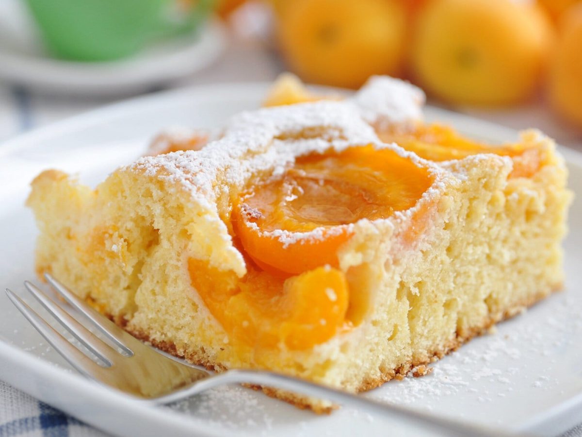 Ein Stück Marillen-Kuchen mit Marzipan vom Blech liegt auf einem weißen Teller. Dahinter liegen einzelne Marillen.