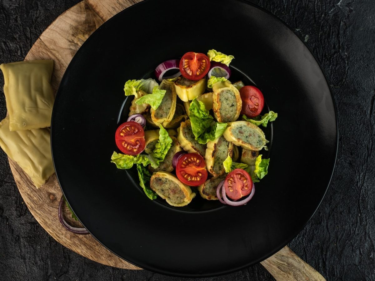 Ein schwarzer Teller mit dem Maultaschen-Salat auf einem Holzbrett und schwarzen Untergrund.