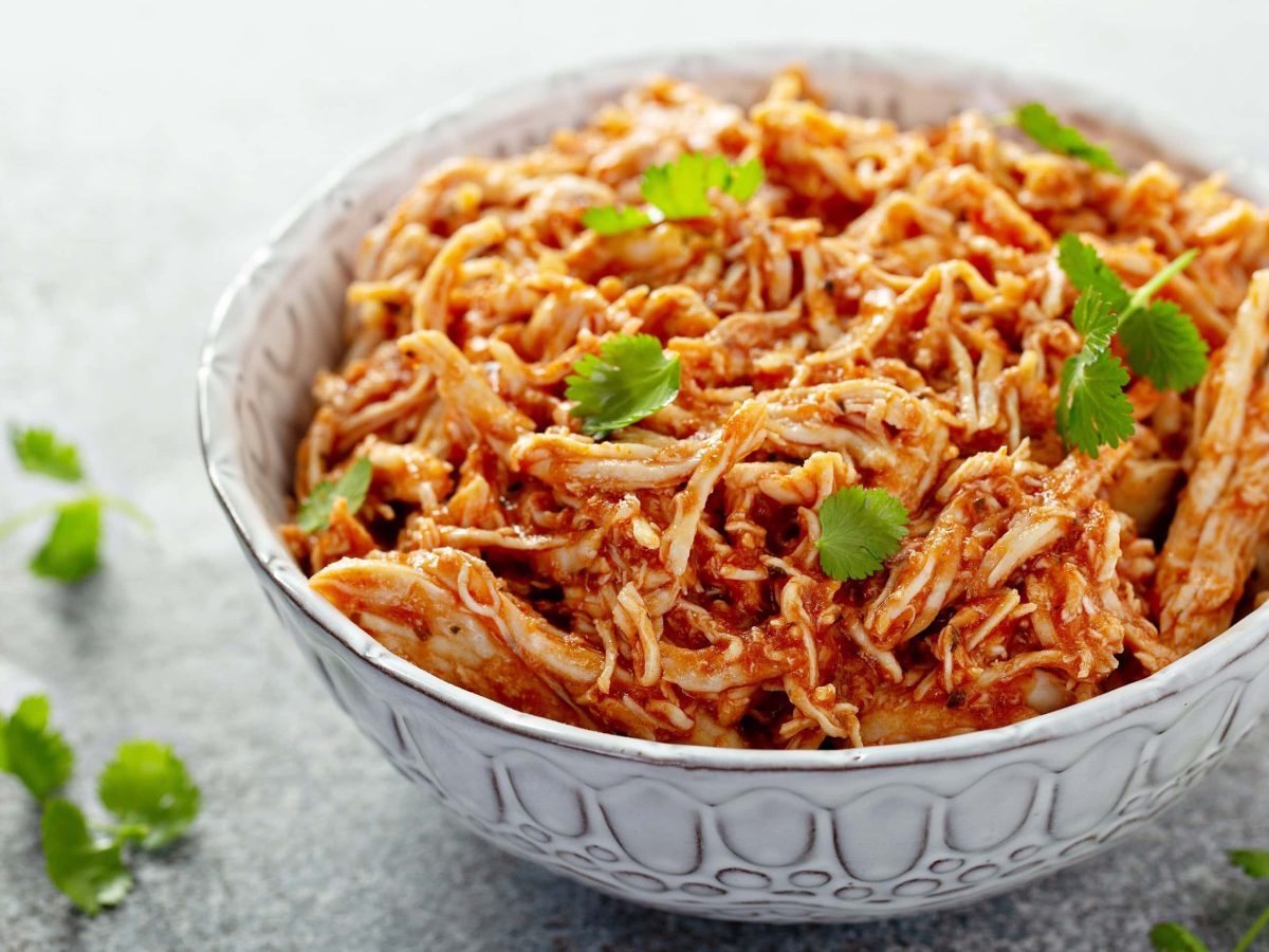 Eine weiße Schüssel mit dem Pulled Chicken auf einem grauen Tisch mit einigen Korianderstielen.