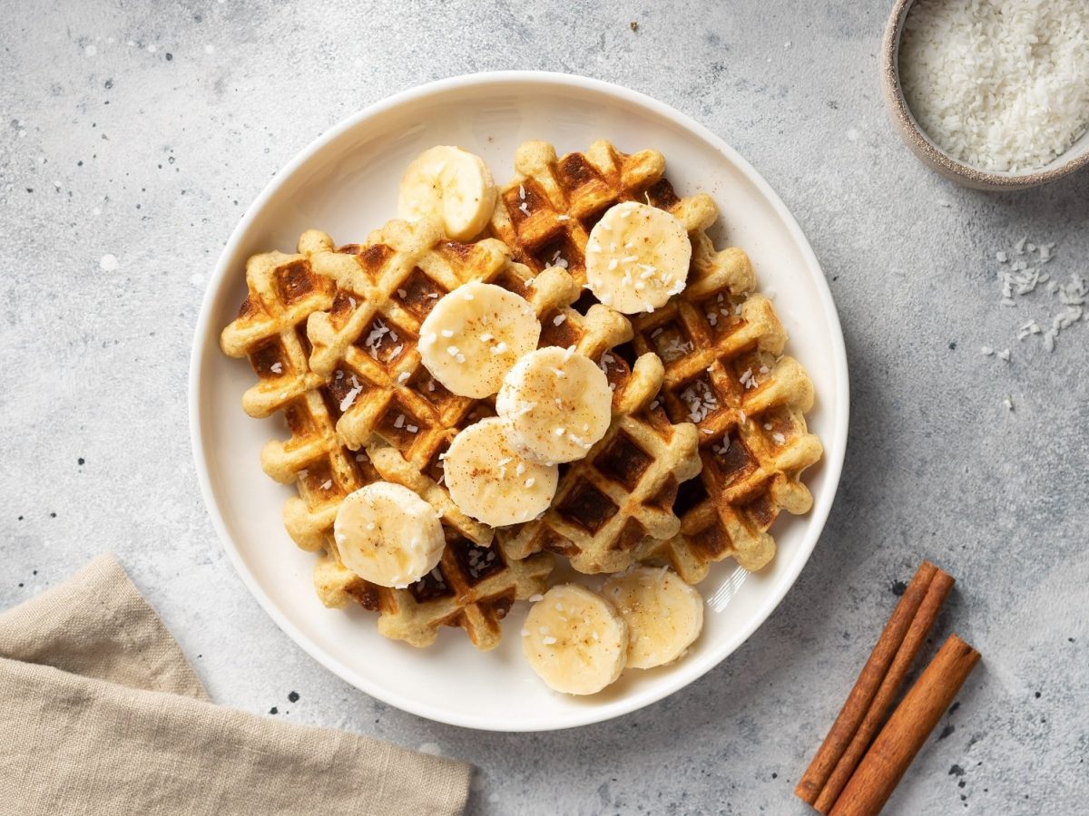 Milchreiswaffeln auf einem Teller mit Bananenscheiben und Kokos, daneben eine Schale Kokosraspeln, eine Serviette und Zimtstangen, alles Draufsicht.