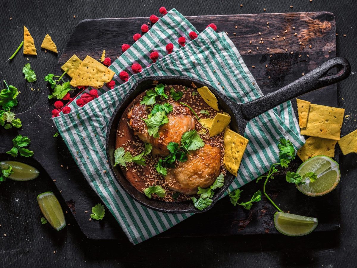 Mexikanisches mole poblano con Pollo, Hähnchenfleisch mit Schokoladensauce, in einer gusseisernen Pfanne, mit Nachos, Koriander und Sesam, Draufsicht.