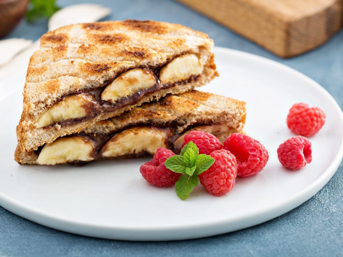 Ein angeschnittenes Bananen-Nutella-Toast auf einem weißen Teller mit frischen Himbeeren.