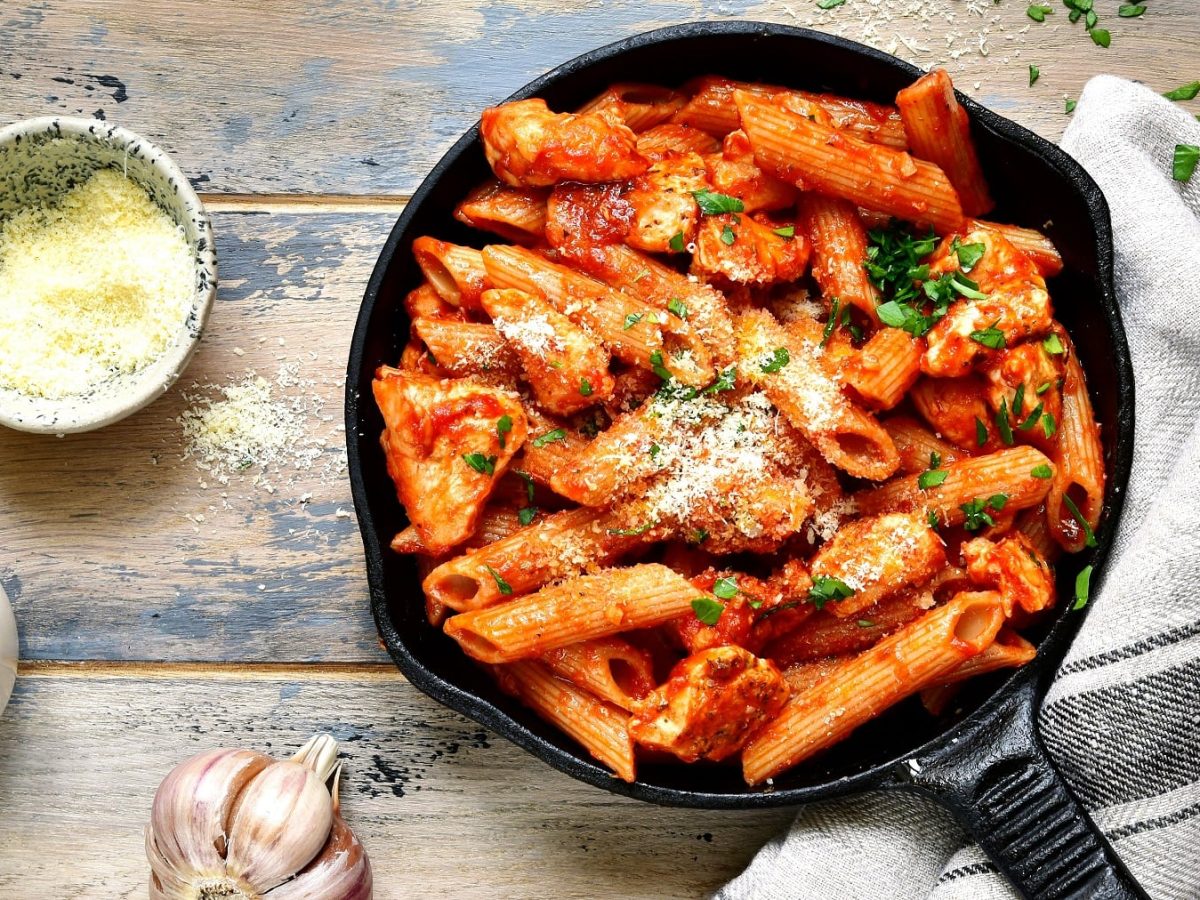 Eine schwarze gusseiserne Pfanne mit der One-Pot-Pasta auf einem grau-blauem Holztisch. Daneben eine Schale mit Parmesan sowie Salz und frischer Knoblauch.