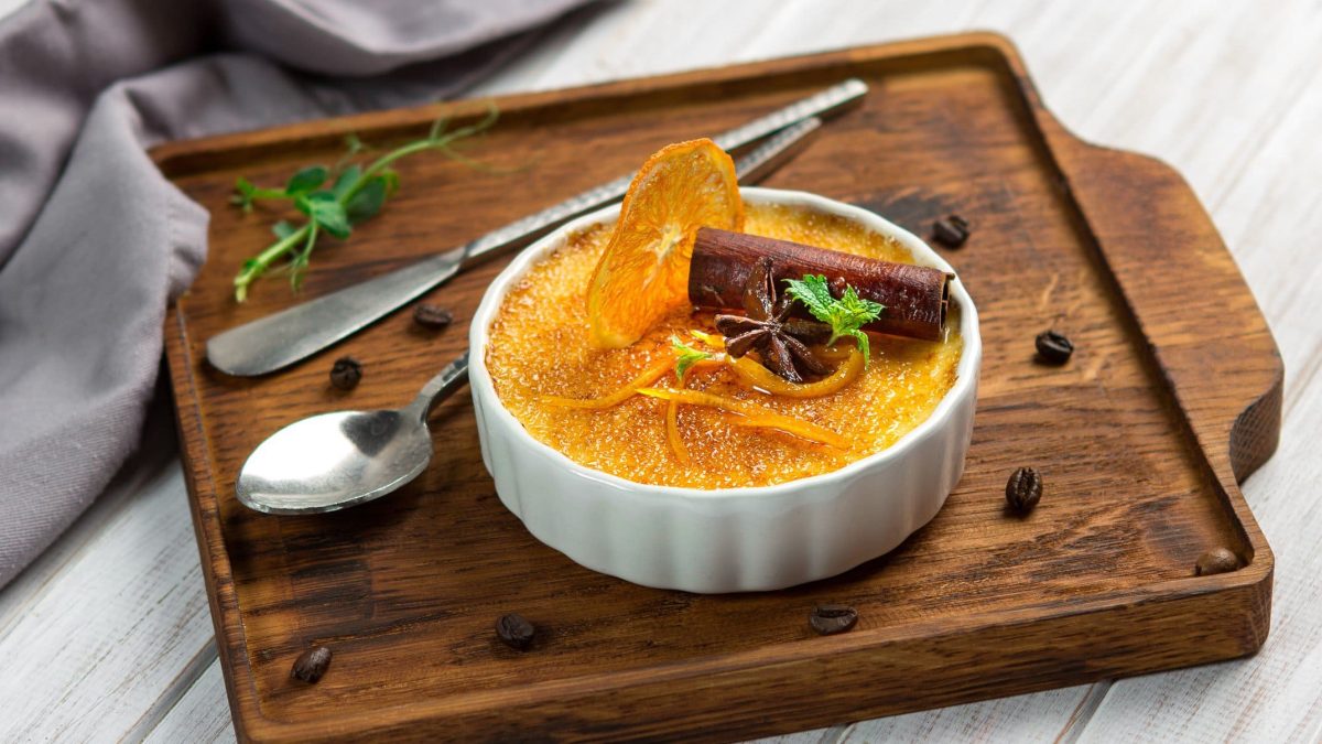 Orangen Crème brûlée mit Zimt, Minze und kandierter Orange in weißer Schale auf Holz auf hellem Untergrund. Im Bild ein Dessertlöffel, Kaffeebohnen und Minze. Draufsicht.