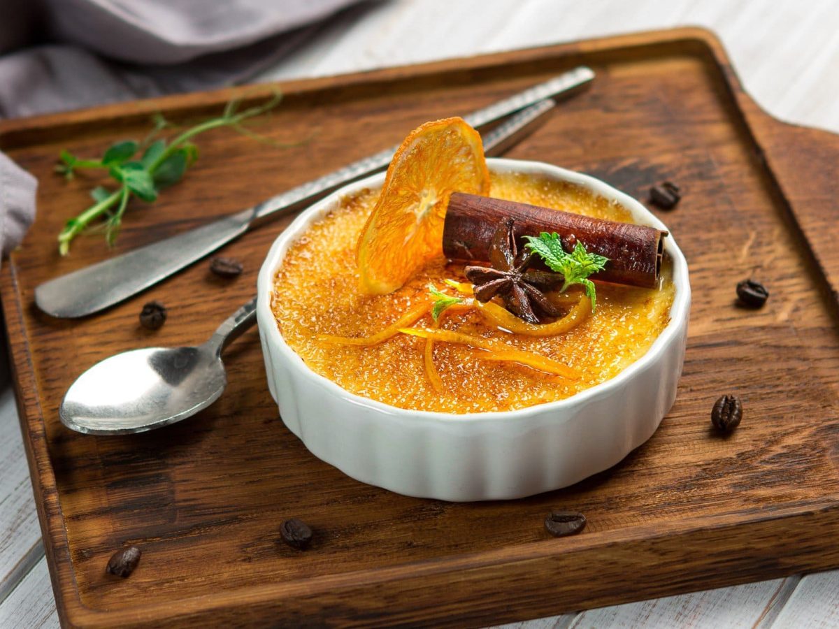 Orangen Crème brûlée mit Zimt, Minze und kandierter Orange in weißer Schale auf Holz auf hellem Untergrund. Im Bild ein Dessertlöffel, Kaffeebohnen und Minze. Draufsicht.