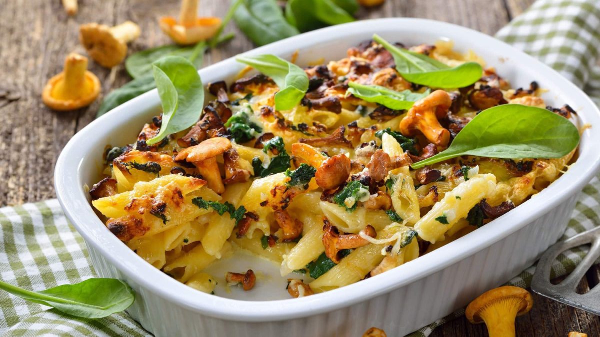 Frontal: Ein Vegetarischer Pasta-Auflauf mit Pfifferlingen steht auf einem Untergrund aus Holz. Daneben liegt ein grün-weiß kariertes Küchenhandtuch