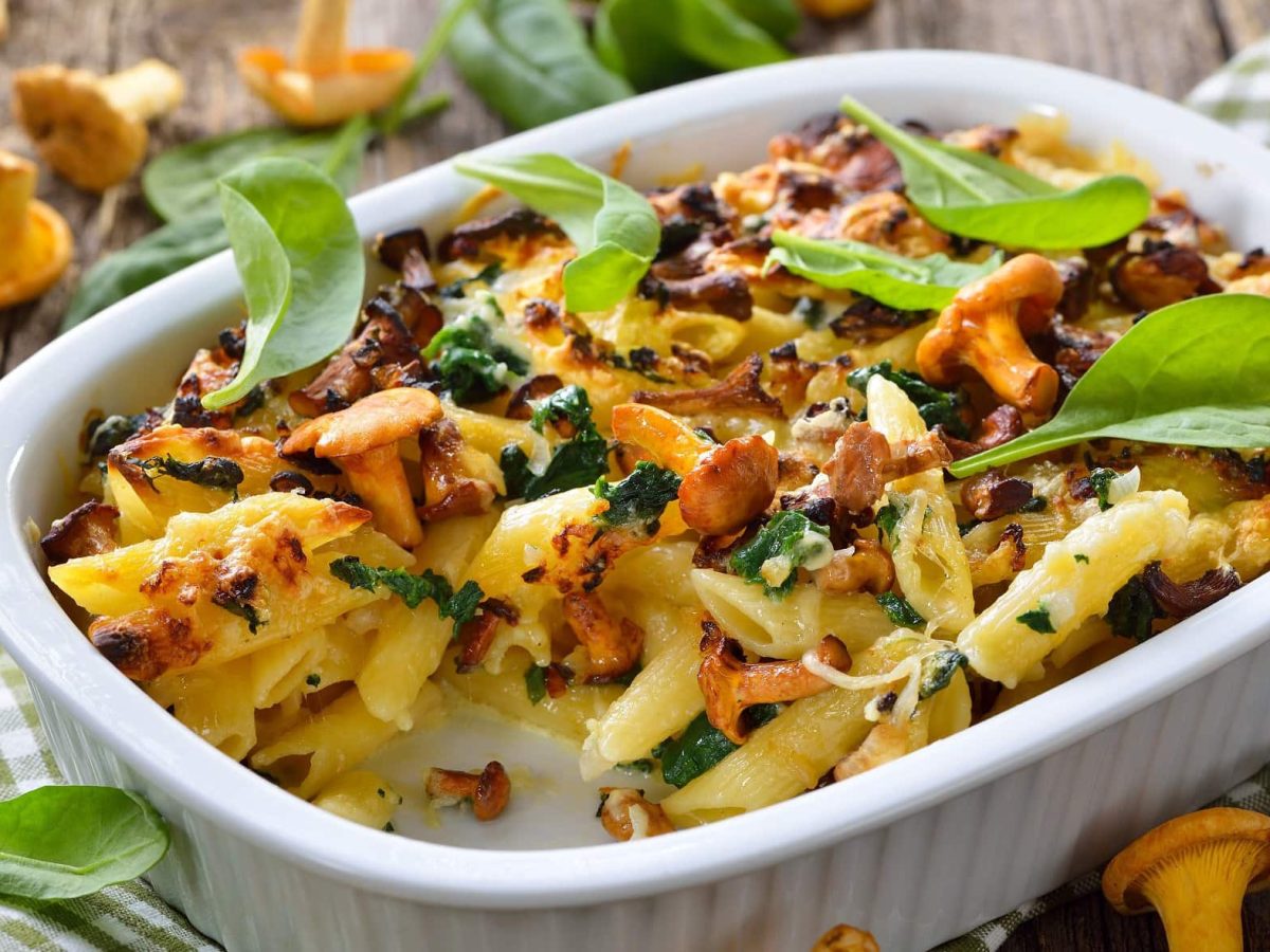 Frontal: Ein Vegetarischer Pasta-Auflauf mit Pfifferlingen steht auf einem Untergrund aus Holz. Daneben liegt ein grün-weiß kariertes Küchenhandtuch