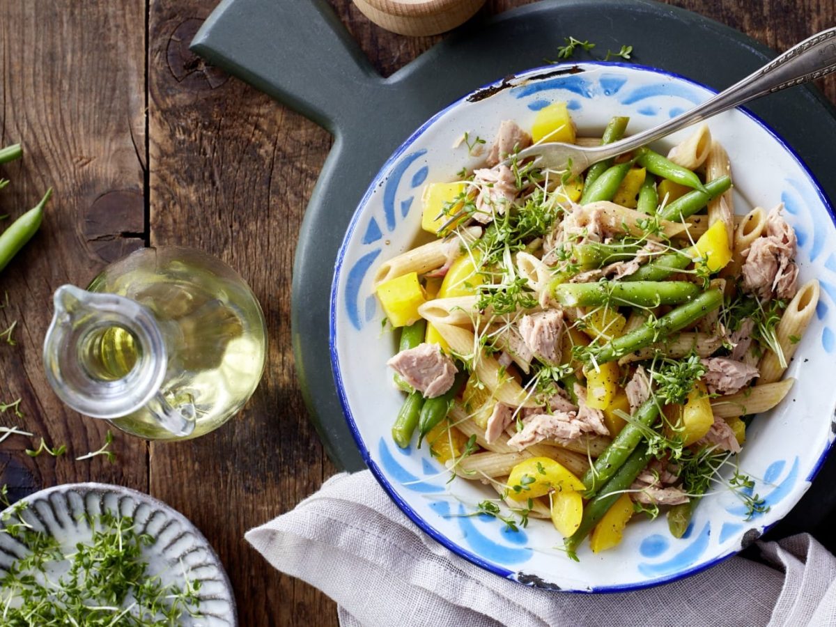 Ein weißer Teller mit blauen Verzierungen gefüllt mit einem Nudelsalat mit Bohnen und Thunfisch auf einem hölzernen Hintergrund. Darunter ein hellgraues Geschirrtuch. Links davon eine kleine Karaffe mit Öl, eine kleine Schüssel mit Kresse und grüne Bohnen.