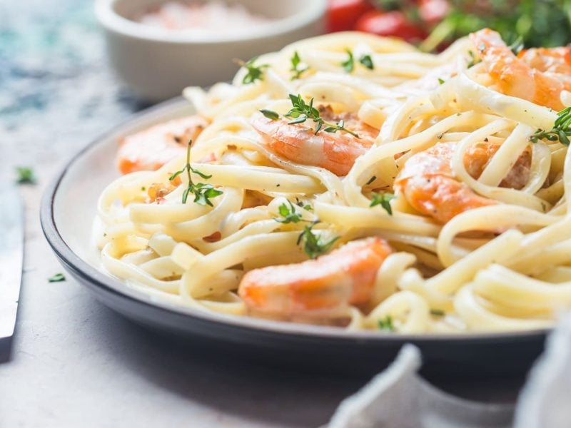 Ein weißer Teller mit blauem Rand und der Pasta mit Lachs und Garnelen auf einem grauen Untergrund sehr nahe fotografiert.