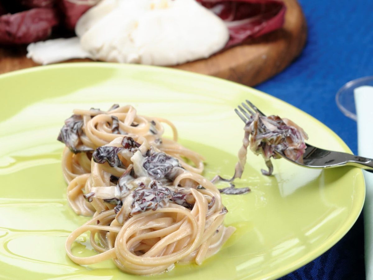 Pasta mit Radicchio und Ziegenfrischkäse auf gelbem Teller mit Gabel. Im Bild Holzbrett mit Radicchio und Ziegenfrischkäse. Draufsicht.