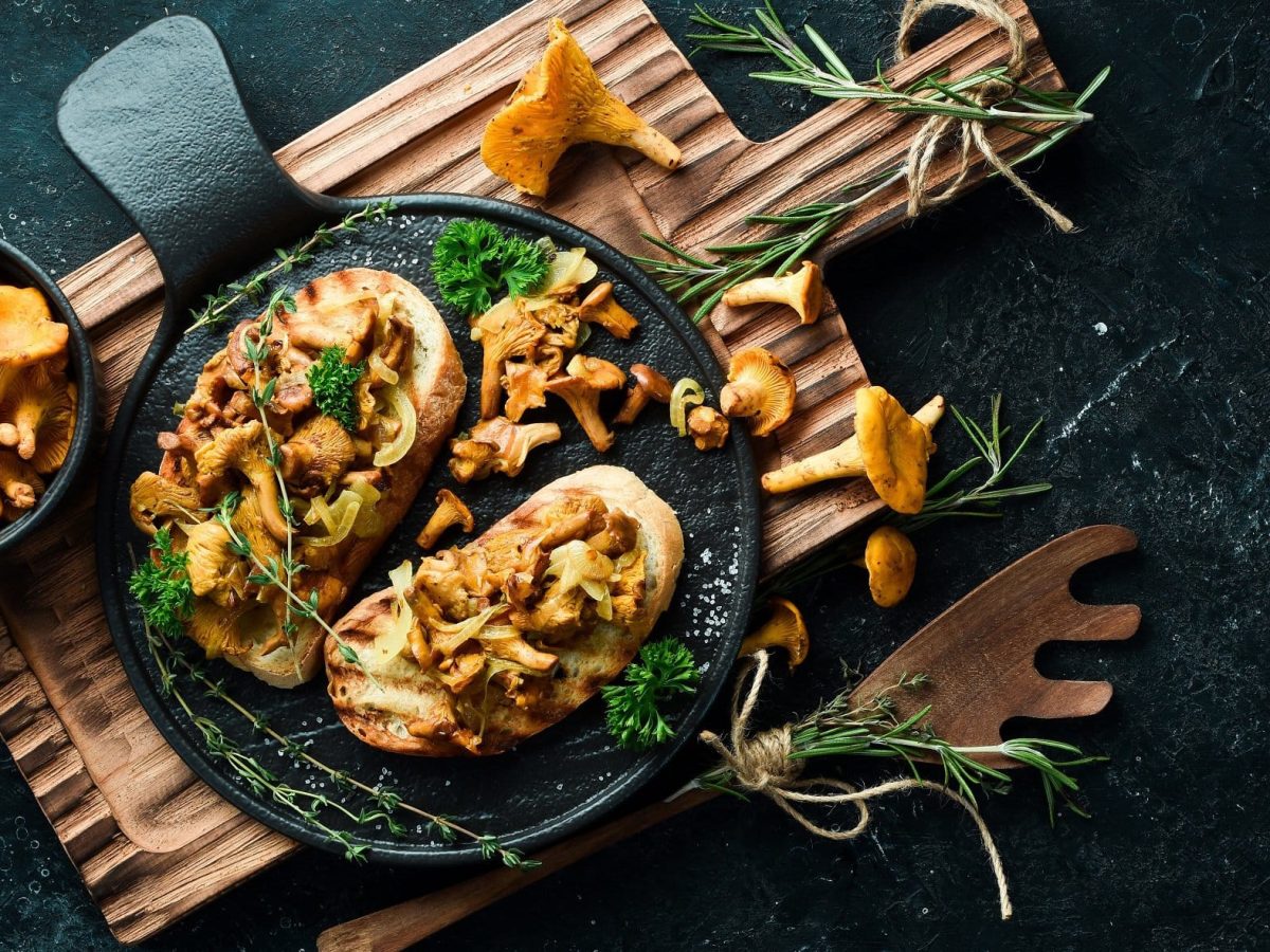 Draufsicht: Dunkler Untergrund, zwei Pecorino-Bruschetta mit Pfifferlingen auf einem Holzbrett und drum herum frische Pfifferlinge und frische Kräuter.