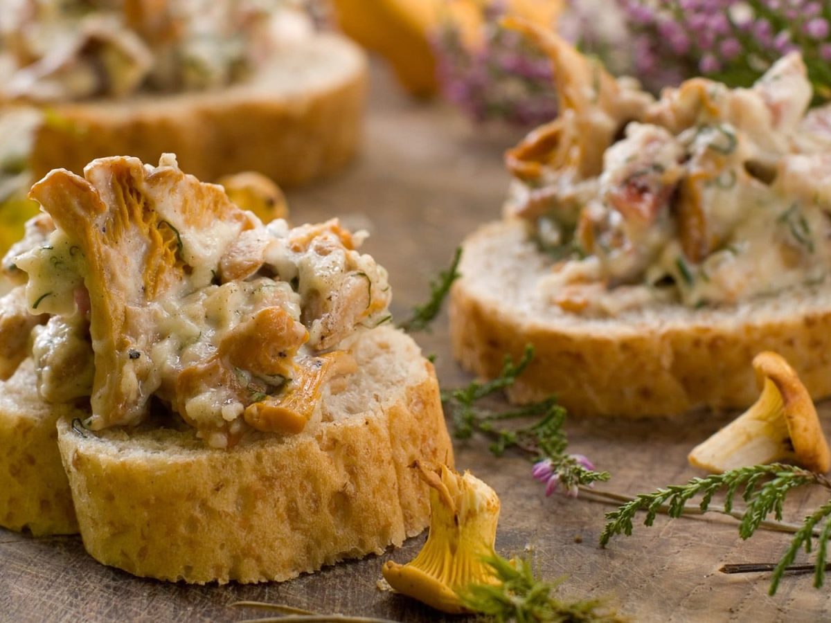 Drei Pfifferling-Häppchen, Baguettescheiben mit gebratenen Rahm-Pfifferlingen belegt, drumherum frische Pilze und blühendes Kraut.