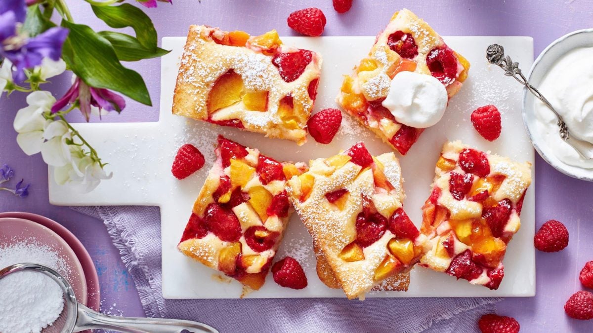 Ein Pfirsich-Melba-Kuchen auf einem weißen Brett auf einem violetten Hintergrund. Links oben Blumen, links unten in einer kleinen Schüssel Puderzucker. Rechts eine Schüssel mit Schlagsahne.