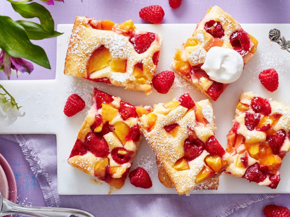 Ein Pfirsich-Melba-Kuchen auf einem weißen Brett auf einem violetten Hintergrund. Links oben Blumen, links unten in einer kleinen Schüssel Puderzucker. Rechts eine Schüssel mit Schlagsahne.