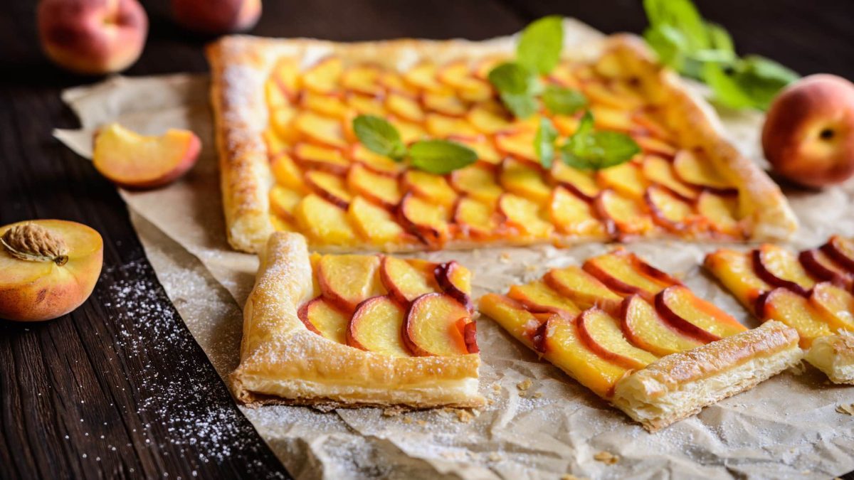 Pfirsich Pie mit Vanillecreme und Blätterteig auf Backpapier auf dunklem Holz. IM Bild weitere Pfirsiche und Minze. Draufsicht.