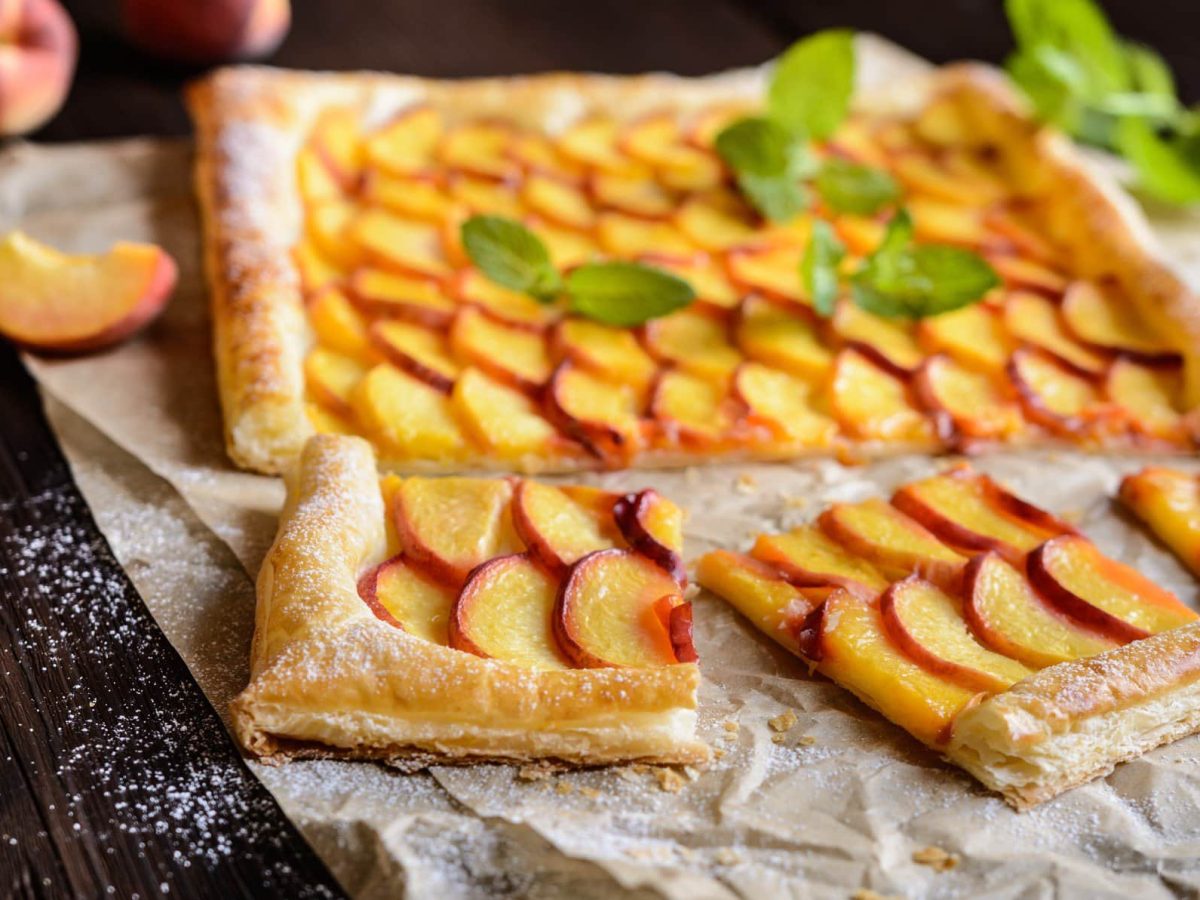 Pfirsich Pie mit Vanillecreme und Blätterteig auf Backpapier auf dunklem Holz. IM Bild weitere Pfirsiche und Minze. Draufsicht.