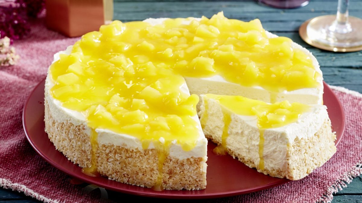 Ein angeschnittener Pina Colada-Cheesecake auf einem roten Teller auf einem roten Geschirrtuch auf einem hölzernen Untergrund. Rechts oben die untere Hälfte von einem Sektglas.