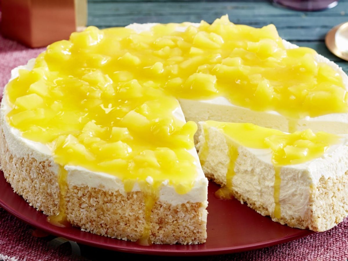 Ein angeschnittener Pina Colada-Cheesecake auf einem roten Teller auf einem roten Geschirrtuch auf einem hölzernen Untergrund. Rechts oben die untere Hälfte von einem Sektglas.