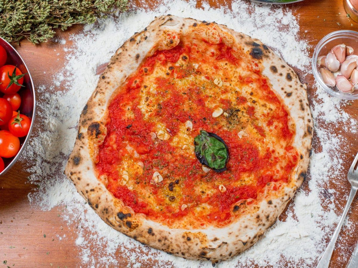 Pizza Marinara in der Draufsicht mit Knoblauch und Basilikum, daneben frische Tomaten in einer Schale, Besteck , getrockneter Oregano, eine Schale Knoblauch, eine mit Basilikum und eine Ölflasche.