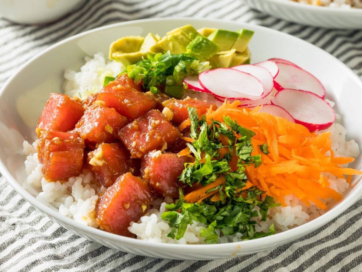 Eine weiße Schüssel mit der Poke Bowl auf einer weiß-grau gestreiften Tischdecke.